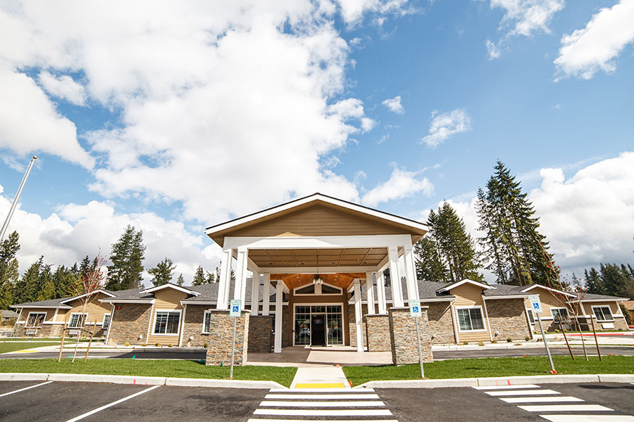 The image shows a modern, welcoming building with a pitched roof and stone accents, surrounded by green landscaping and trees, under a partly cloudy sky.