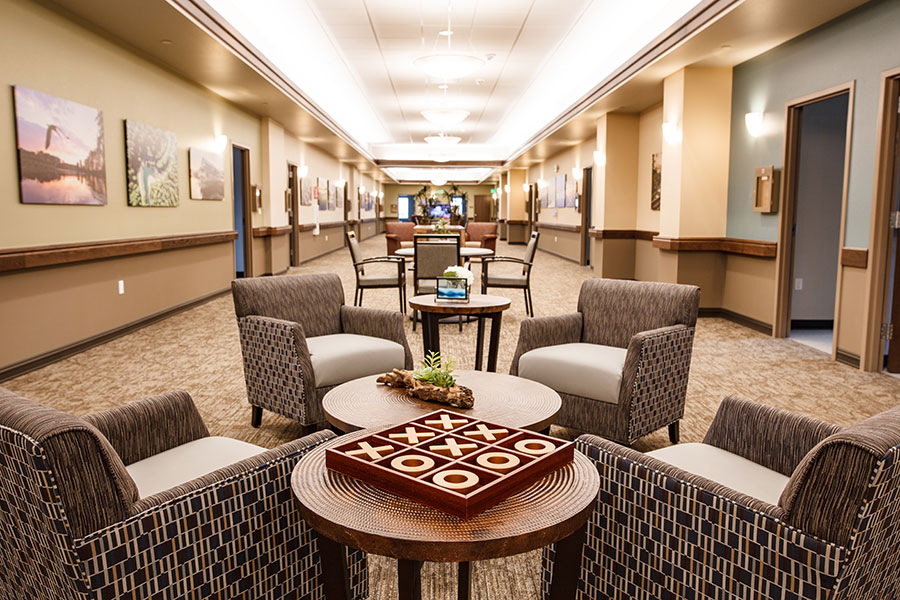 The image depicts a modern, inviting hallway with seating areas. It features comfortable chairs grouped around small tables, adorned with decor such as plants and a tic-tac-toe game. The walls showcase artwork, contributing to a warm, communal atmosphere.