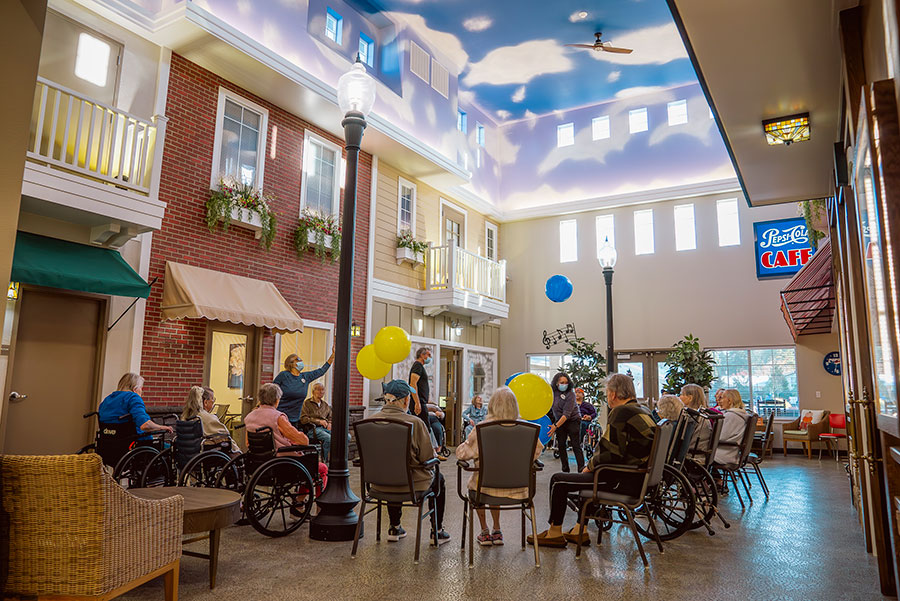 The image shows a lively indoor space resembling a small town square, where a group of elderly individuals in wheelchairs and chairs are participating in activities. There are colorful balloons, greenery, and a painted sky on the ceiling, creating a cheerful atmosphere.