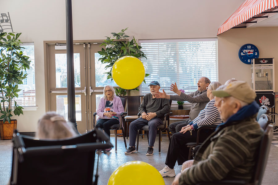 The image depicts a cheerful scene in a communal space where several elderly individuals are interacting, some seated and engaged in conversation, with a large yellow balloon adding a lively touch to the atmosphere.