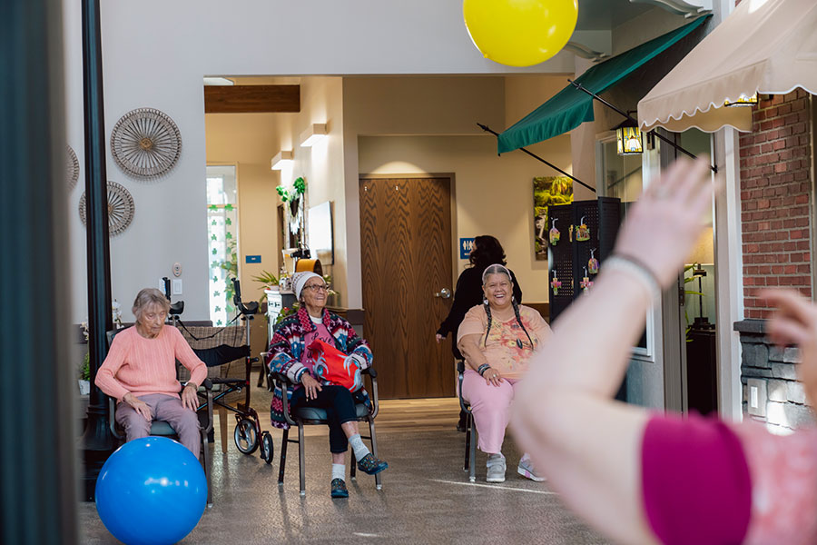 The image depicts a cheerful indoor setting with three elderly women seated, engaged in an activity, while a colorful balloon floats above. Bright lighting and a welcoming atmosphere are evident.