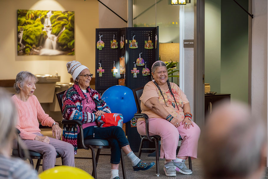 The image shows three elderly women sitting together and smiling in a bright, cheerful indoor setting. There are colorful decorations in the background, and a water feature can be seen in a photo on the wall. The atmosphere appears lively and friendly.