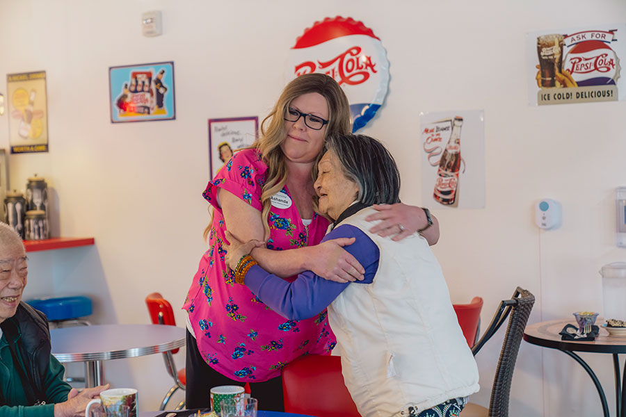 The image shows a warm moment in a retro-themed diner, where a woman in a floral shirt is embracing an elderly woman. A man sits nearby, smiling, creating a friendly and heartfelt atmosphere.