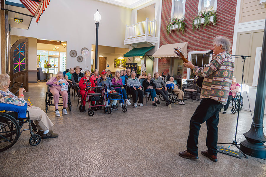 A musician performs for a group of elderly residents seated in wheelchairs and chairs, enjoying a lively gathering in a cozy indoor space.