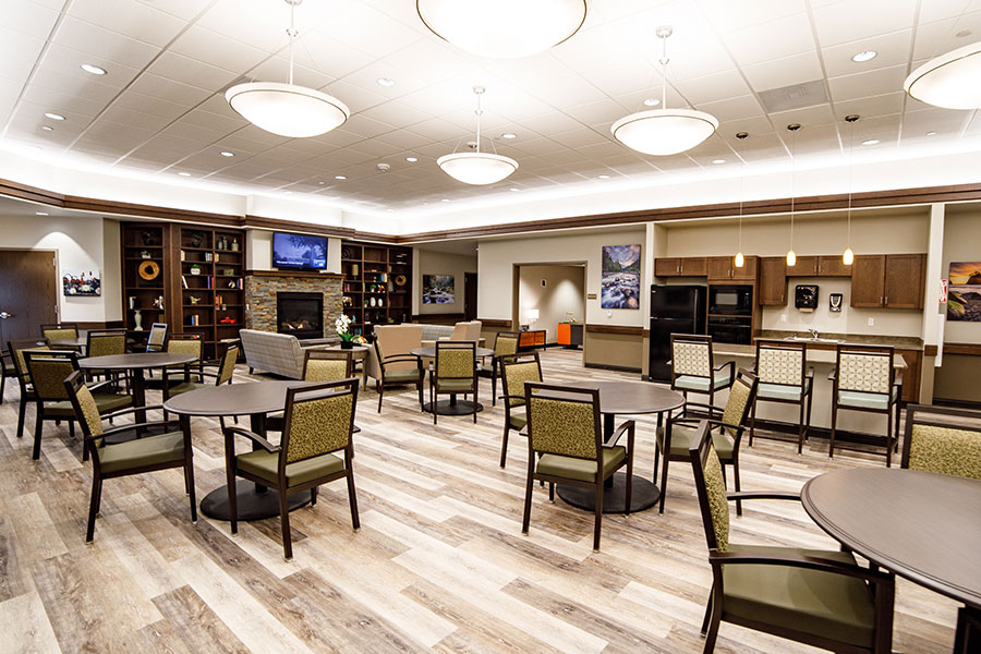 A modern, spacious common area featuring circular tables, chairs, a stone fireplace, and a kitchenette, with warm lighting and a cozy atmosphere.