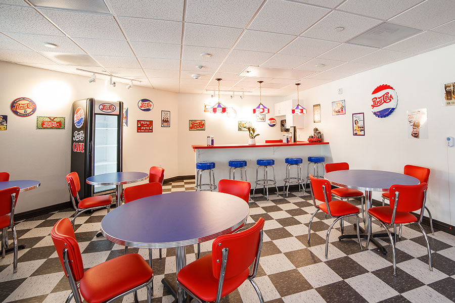 A retro-style diner with red chairs, round tables, and a soda bar featuring vintage Pepsi decor.