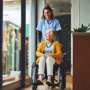 A caregiver pushes an older woman in a wheelchair through a memory care community.