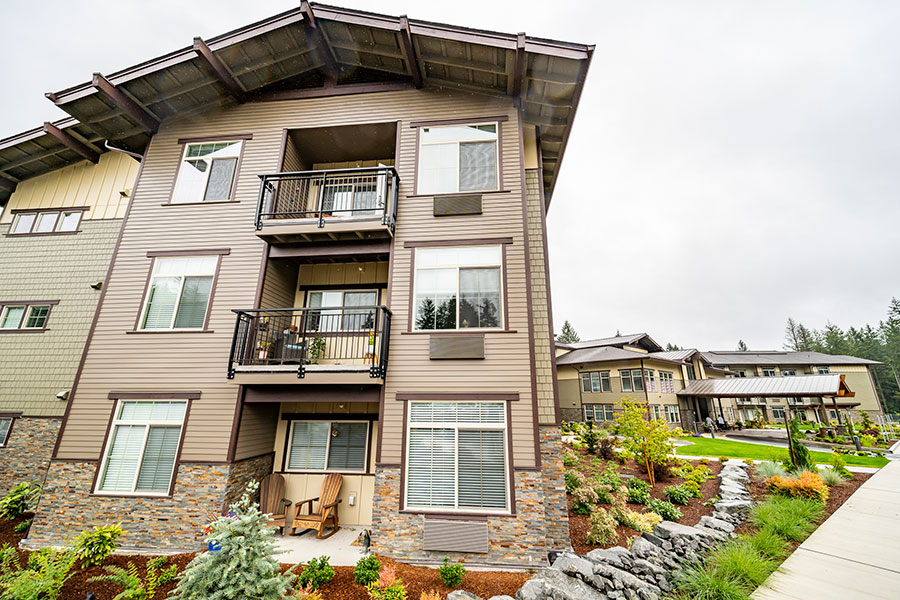 The image shows a multi-story apartment building with modern design elements, featuring balconies, large windows, and stone accents, set amidst landscaped greenery.