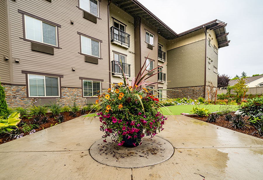 The image features a landscaped area in front of a modern apartment building, with a vibrant flower pot at the center, surrounded by greenery and paved walkways.