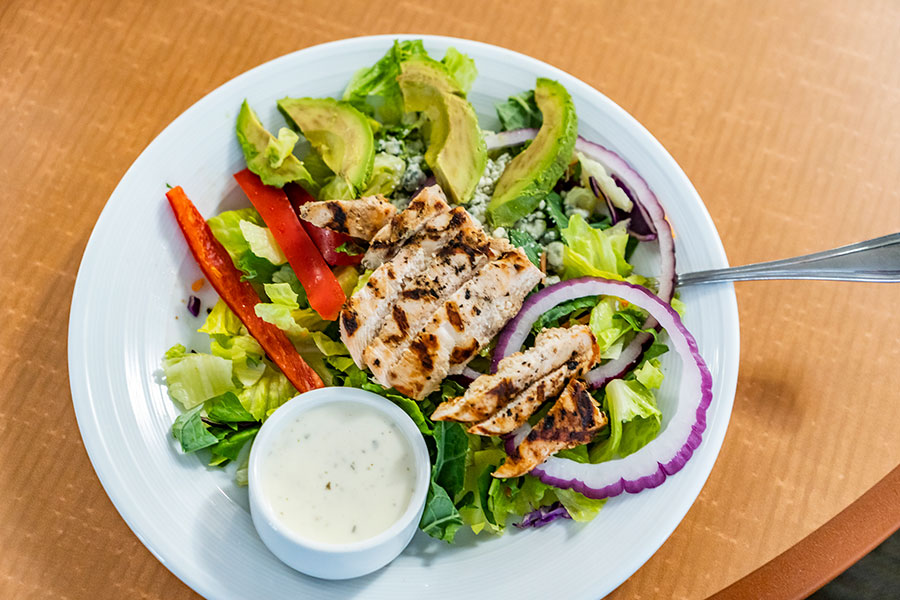 A vibrant salad with grilled chicken, avocado, red bell peppers, onion, and a side of creamy dressing.