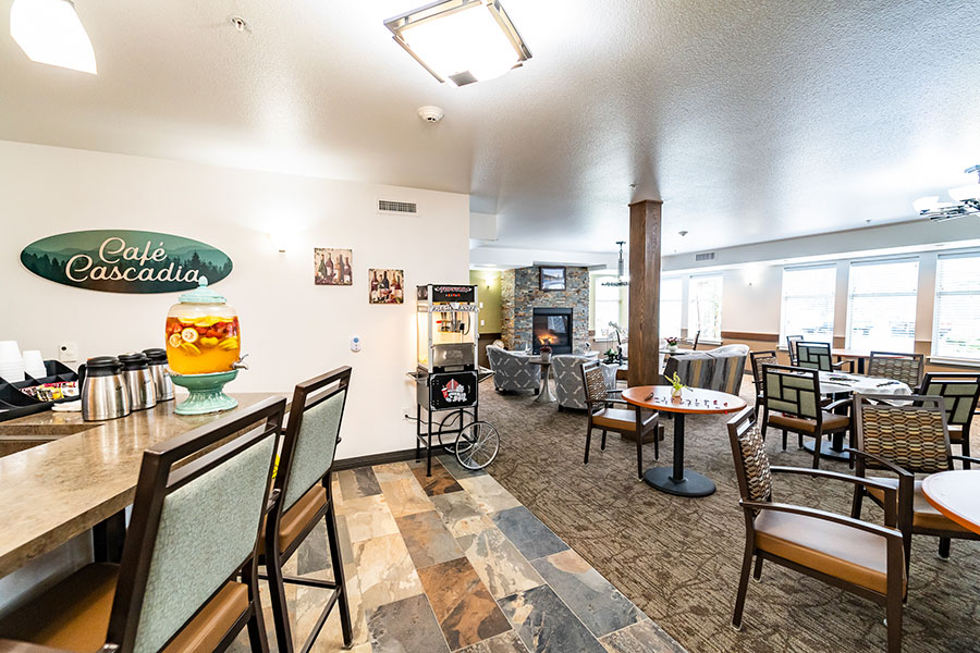 A cozy café interior featuring a sign for "Café Cascadia," a drink dispenser with fruit, and seating arranged around tables. There's a fireplace and large windows for natural light.