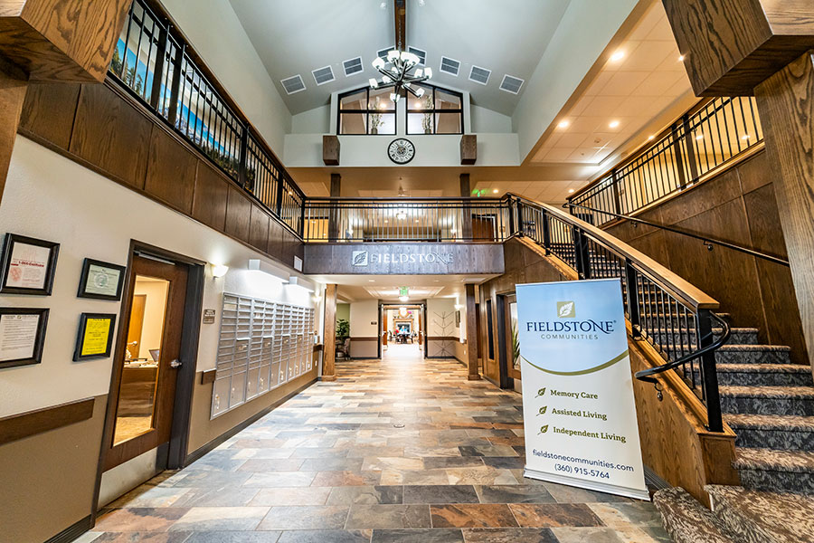 The image depicts a welcoming lobby of a facility, featuring a staircase, a reception area, and signage for Fieldstone Communities, emphasizing services like memory care and assisted living. The space has a warm design with wooden accents and a high ceiling.