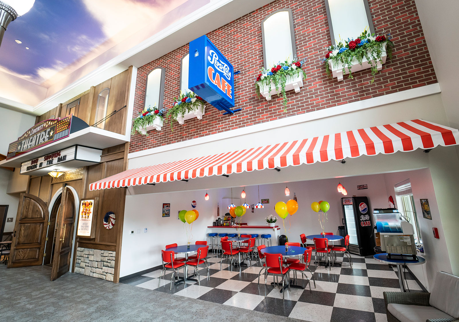 The image depicts a vibrant café interior with a retro design, featuring red and white striped awnings, colorful balloons, and diner-style seating. The space also includes a theater entrance and decorative wall features, creating a lively atmosphere.