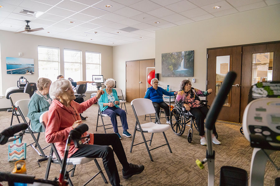 The image shows a group of elderly individuals sitting in a brightly lit room, participating in a seated activity, likely involving exercise or relaxation. Some are using mobility aids, and there are fitness machines in the background. The atmosphere appears engaging and supportive.