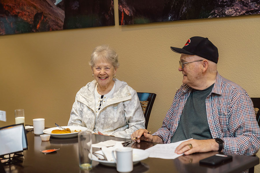 Two older adults, seated at a table with food and drinks, enjoying a conversation and smiling.