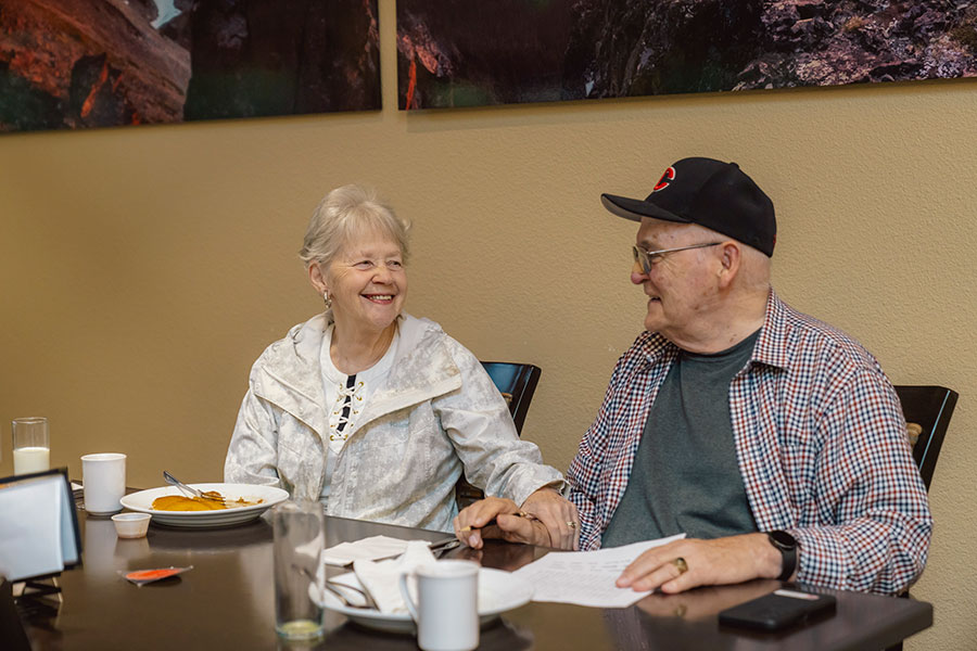 An elderly couple shares a warm moment over breakfast, smiling and holding hands at a table with food and drinks. The environment appears cozy and inviting.