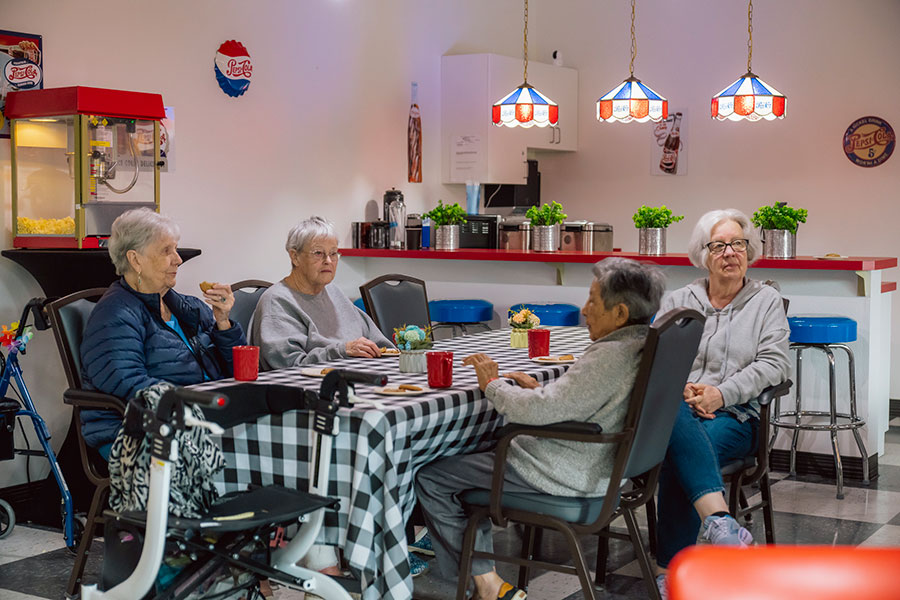 A cozy dining area with four elderly women seated around a checkered table, enjoying snacks and drinks in a retro-style cafe setting.