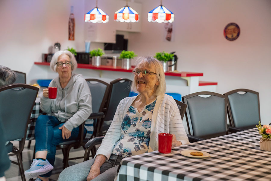 The image shows two elderly women sitting in a cozy, well-lit room with a checkered tablecloth, smiling and enjoying their time. One is holding a red cup, and there is a plate with a cookie on the table.