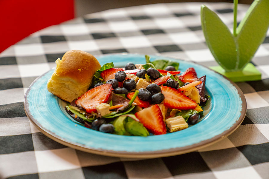 A colorful salad with strawberries, blueberries, and greens, served with a small roll on a vibrant blue plate, placed on a black-and-white checkered tablecloth.